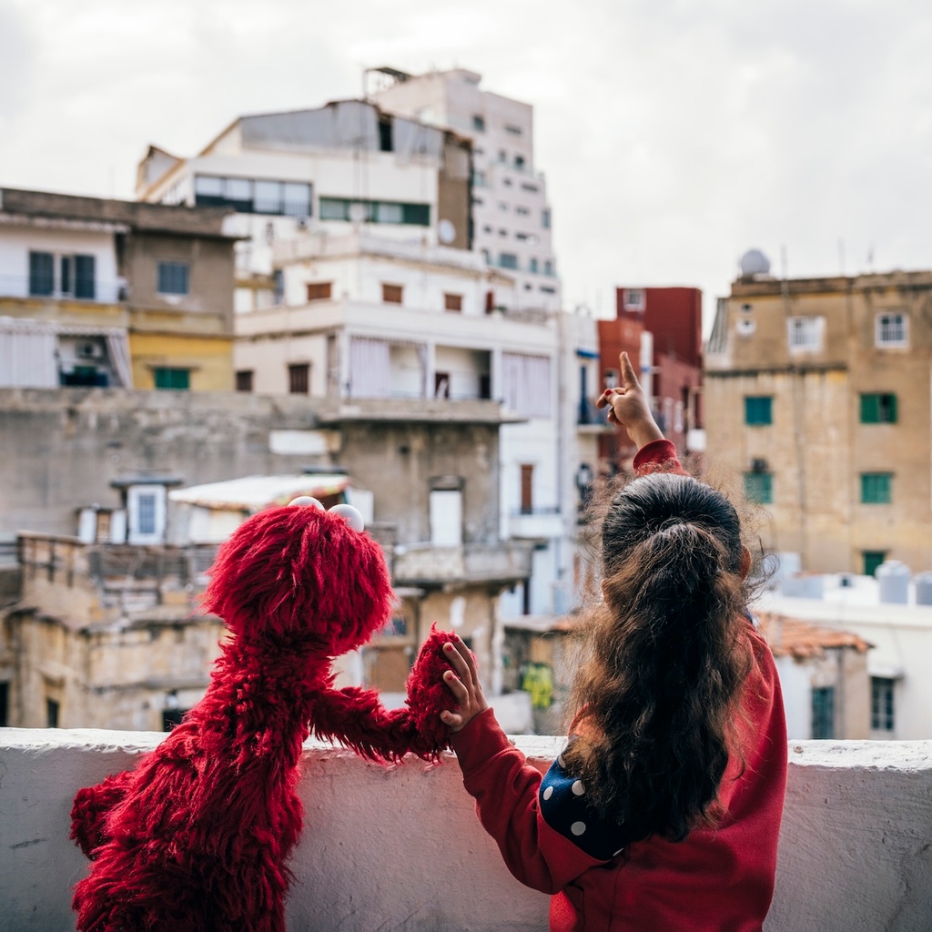 Elmo from Sesame Street and a young girl hold hands and look out from a balcony in a war torn city